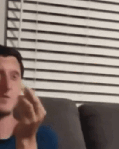 A Man Is Sitting On A Couch In Front Of A Window With Blinds And Holding Something In His Hand . GIF