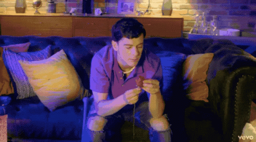 A Man Is Sitting On A Couch Looking At His Phone With The Word Vevo On The Bottom Left GIF