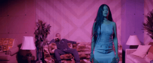 A Man Is Sitting On A Couch While A Woman In A Blue Dress Stands In Front Of Him . GIF