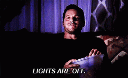a man is sitting on a couch with a bucket of popcorn and the words lights are off above him