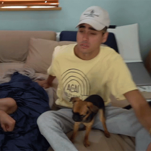 A Man Is Sitting On A Couch With A Small Dog Wearing A Yellow Shirt That Says Agai GIF