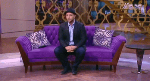 A Man Is Sitting On A Purple Couch With A Hat On GIF