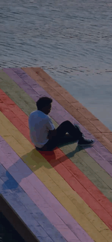 A Man Is Sitting On A Rainbow Colored Dock Overlooking A Body Of Water GIF