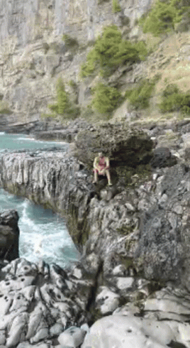 A Man Is Sitting On A Rock Near The Water . GIF