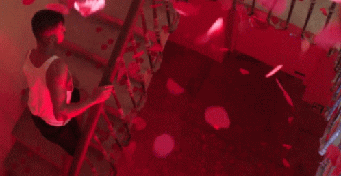 A Man Is Sitting On A Set Of Stairs With Petals Falling From The Ceiling . GIF