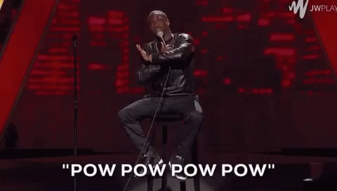 A Man Is Sitting On A Stool On A Stage Holding A Microphone And Talking Into It . GIF