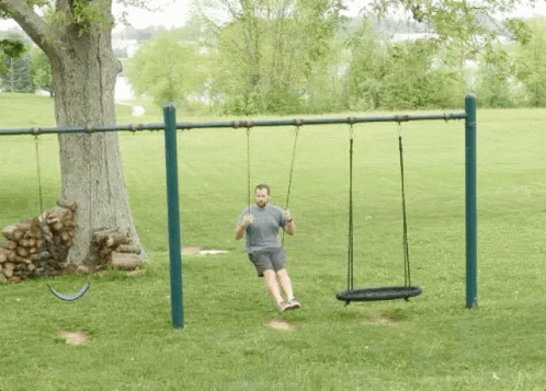 A Man Is Sitting On A Swing In A Park . GIF