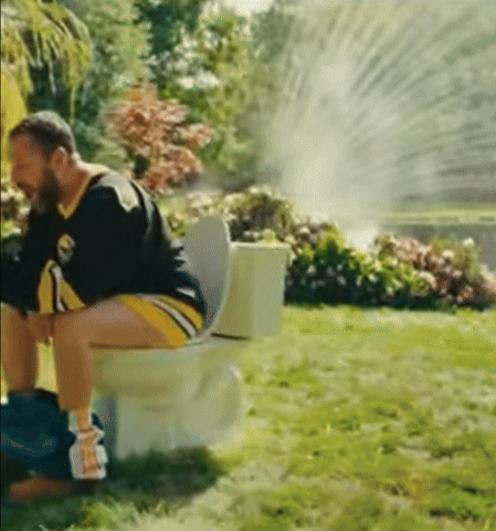 A Man Is Sitting On A Toilet In The Grass With A Sprinkler Spraying Water GIF
