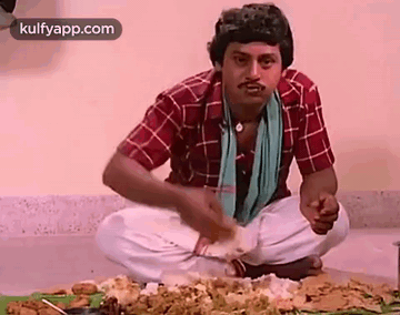 A Man Is Sitting On The Floor Eating Food From A Table . GIF