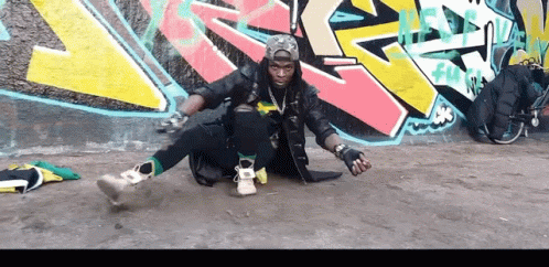 A Man Is Sitting On The Ground In Front Of A Wall With Graffiti On It That Says ' Fun ' GIF
