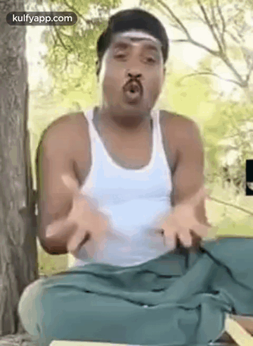 A Man Is Sitting Under A Tree With His Hands Outstretched And Making A Funny Face . GIF