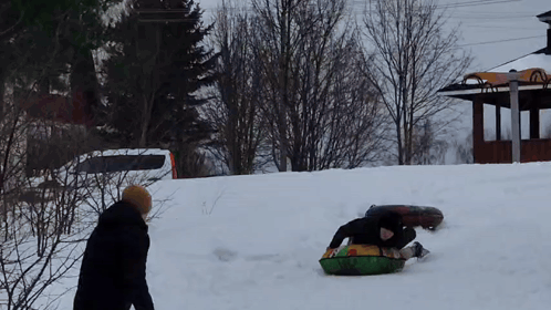 A Man Is Sledding Down A Snow Covered Hill While Another Man Watches GIF