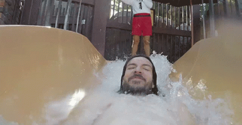 A Man Is Sliding Down A Water Slide With A Lifeguard Standing Behind Him GIF