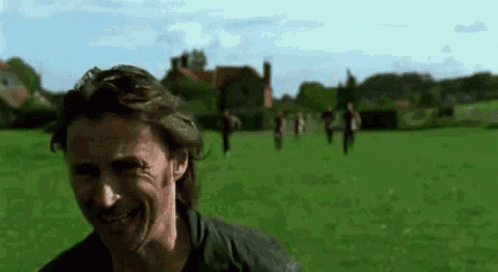 A Man Is Smiling In A Field With A Group Of People Walking In The Background . GIF