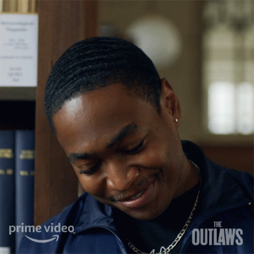 A Man Is Smiling In Front Of A Bookshelf With The Outlaws Written On The Bottom Right Corner GIF