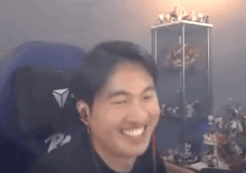 A Man Is Smiling While Sitting In A Chair In Front Of A Display Case Of Figurines . GIF