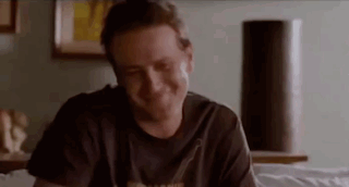A Man Is Smiling While Sitting On A Bed In A Room . GIF