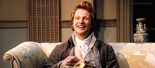 A Man Is Smiling While Sitting On A Couch With A Vase In The Background .. GIF