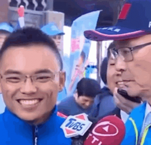 A Man Is Smiling While Talking Into A Microphone While Wearing Glasses And A Hat . GIF