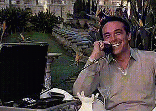 A Man Is Smiling While Talking On A Phone Next To A Briefcase GIF