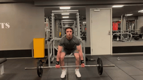 A Man Is Squatting Down Holding A Barbell In A Gym With The Word Ts On The Wall GIF