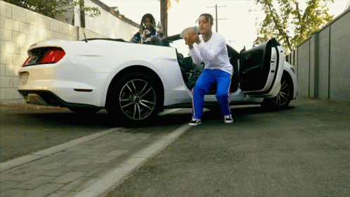 A Man Is Squatting In Front Of A White Car Holding A Bag Of Money GIF