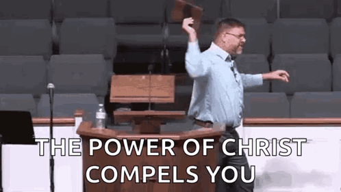 A Man Is Standing At A Podium In Front Of A Microphone Holding A Bible In His Hand . GIF