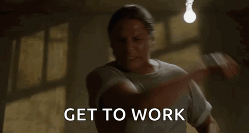 A Man Is Standing In A Dark Room With A Light Bulb Hanging From The Ceiling And Says `` Get To Work '' . GIF