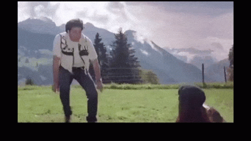 A Man Is Standing In A Field With Mountains In The Background While A Woman Looks On GIF
