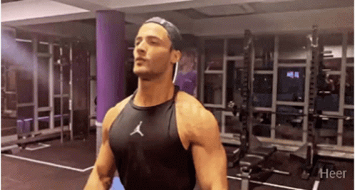 A Man Is Standing In A Gym Wearing A Black Tank Top And A Hat . GIF