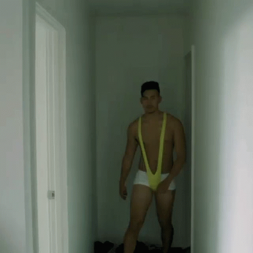 A Man Is Standing In A Hallway Wearing A Yellow Slingshot Underwear . GIF