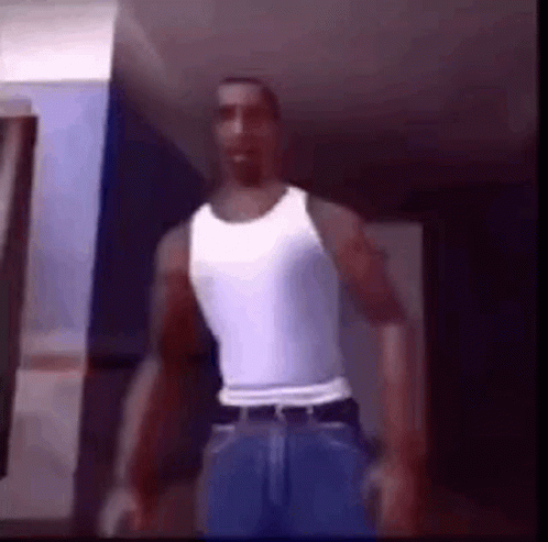 A Man Is Standing In A Room Wearing A White Tank Top And Blue Jeans . GIF