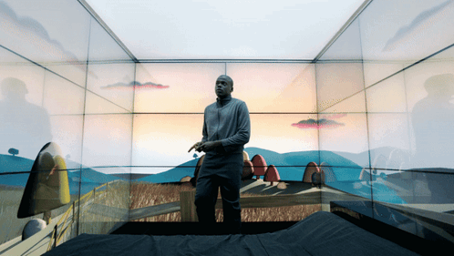 A Man Is Standing In A Room With A Painting Of A Landscape On The Wall GIF