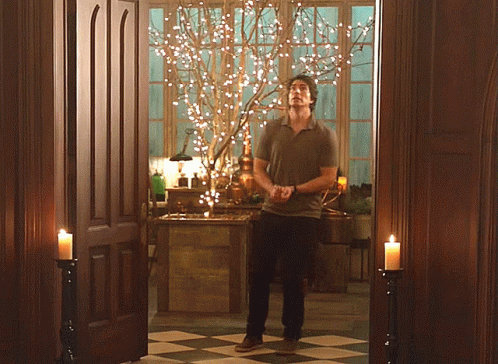 A Man Is Standing In A Room With Candles And A Tree With Lights On It GIF