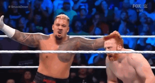 A Man Is Standing In A Wrestling Ring With His Arms Outstretched Next To Another Man . GIF