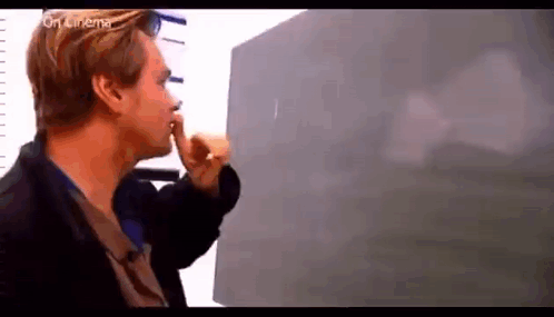 A Man Is Standing In Front Of A Blackboard Holding His Finger To His Lips . GIF