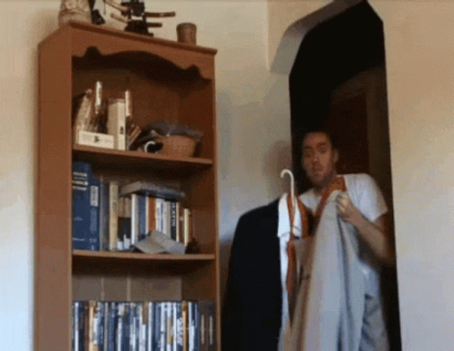 A Man Is Standing In Front Of A Bookshelf With A Book Titled ' A Dictionary ' On The Top Shelf GIF
