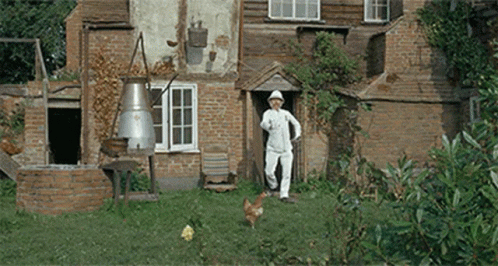 A Man Is Standing In Front Of A Brick House With A Chicken In Front Of Him GIF