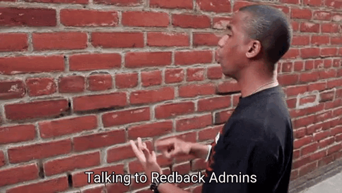 A Man Is Standing In Front Of A Brick Wall Talking To Redback Admins GIF