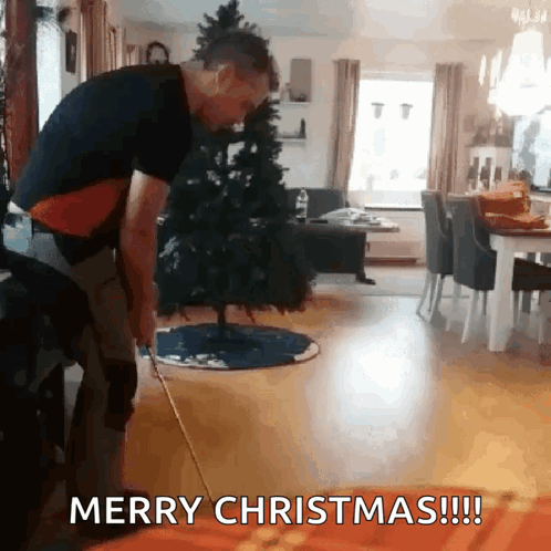 A Man Is Standing In Front Of A Christmas Tree With The Words Merry Christmas Written On The Bottom GIF