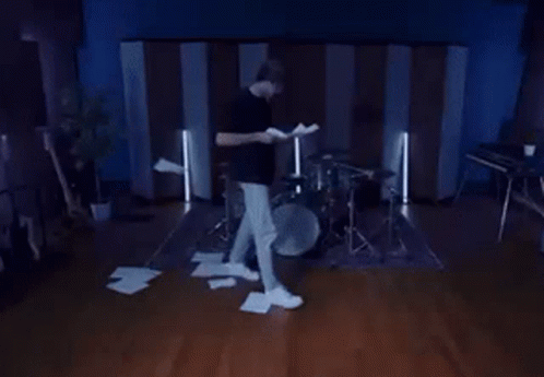A Man Is Standing In Front Of A Drum Set Holding A Piece Of Paper GIF
