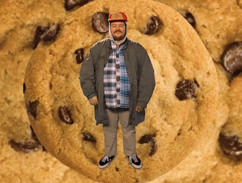 A Man Is Standing In Front Of A Large Cookie With Chocolate Chips GIF