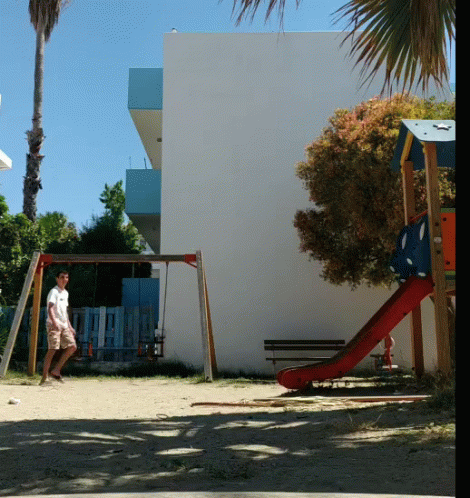 A Man Is Standing In Front Of A Playground With A Slide And Swings GIF