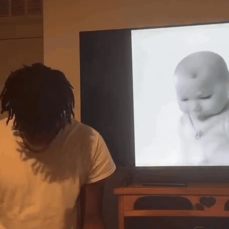 A Man Is Standing In Front Of A Television With A Baby On The Screen GIF