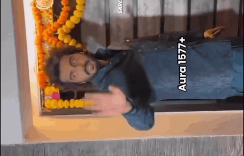 A Man Is Standing In Front Of A Wall With Flowers And A Sign That Says Aura 1577+ On It GIF