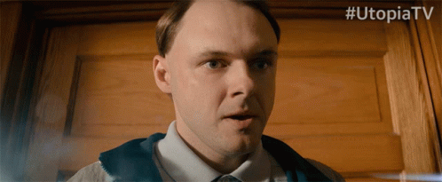 A Man Is Standing In Front Of A Wooden Door With # Utopiatv Written Above Him GIF