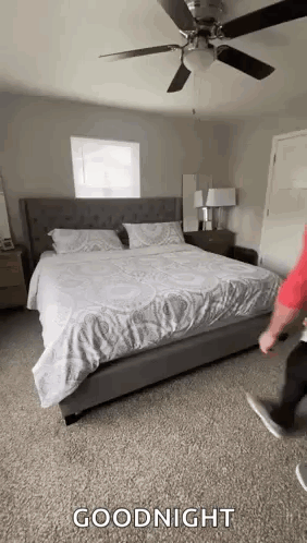 a man is standing next to a bed in a bedroom with a ceiling fan and a window .