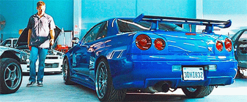 A Man Is Standing Next To A Blue Car With A License Plate That Says Bdh1832 . GIF