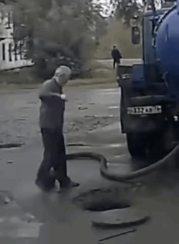 A Man Is Standing Next To A Blue Truck That Is Pumping Water Into A Manhole . GIF