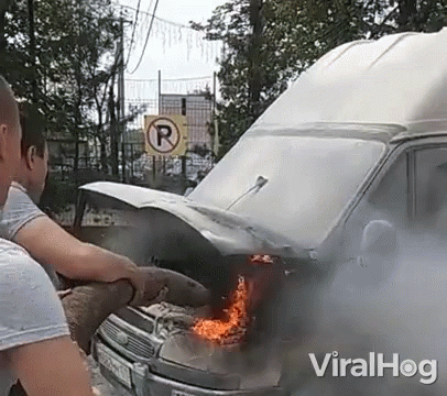 A Man Is Standing Next To A Burning Van With Smoke Coming Out Of It . GIF